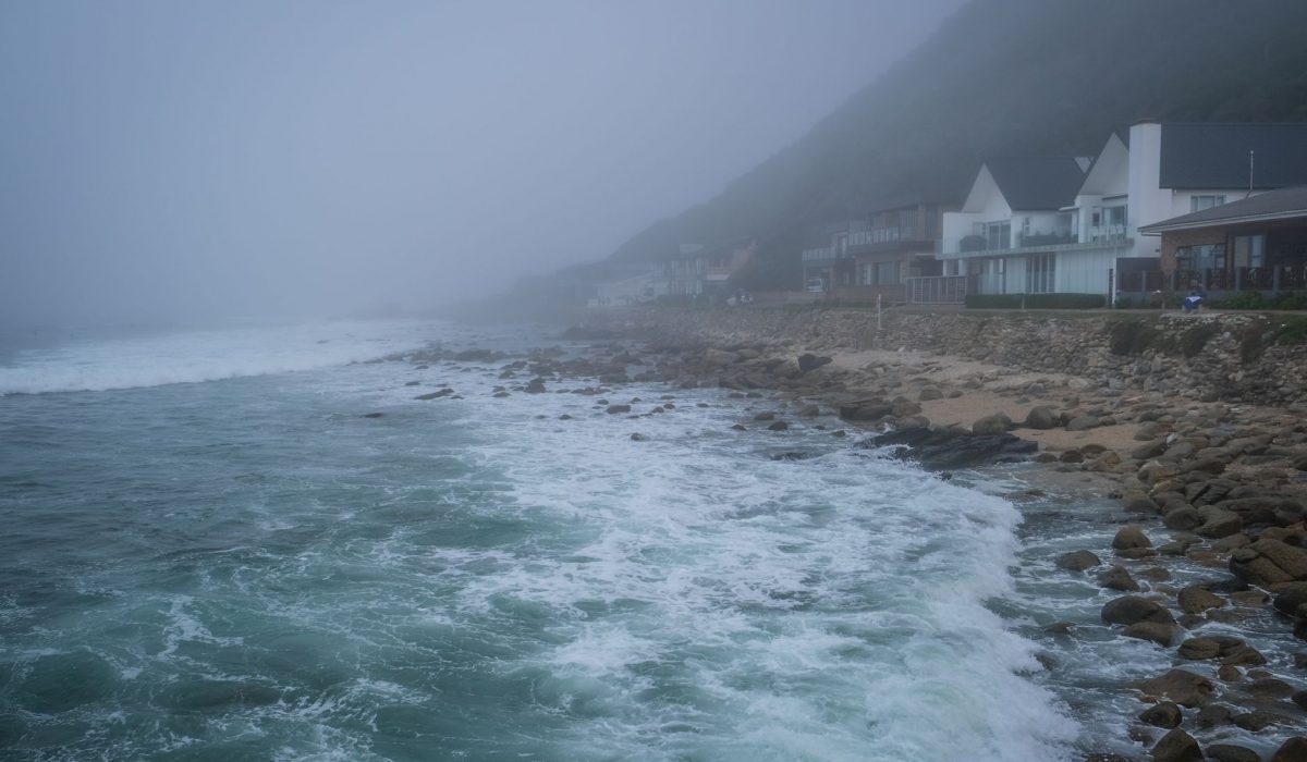 A foggy coastline with waves hitting the rocky shore and modern houses in view
