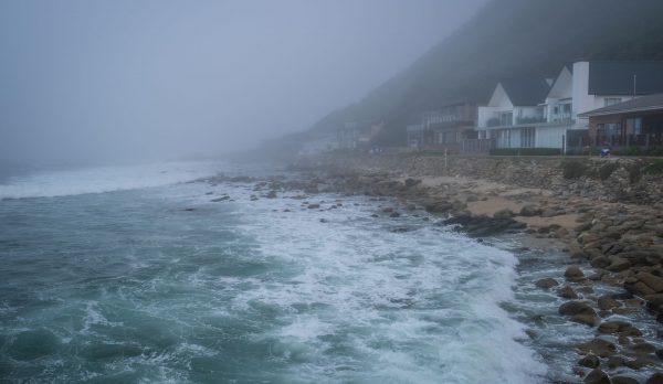 A foggy coastline with waves hitting the rocky shore and modern houses in view